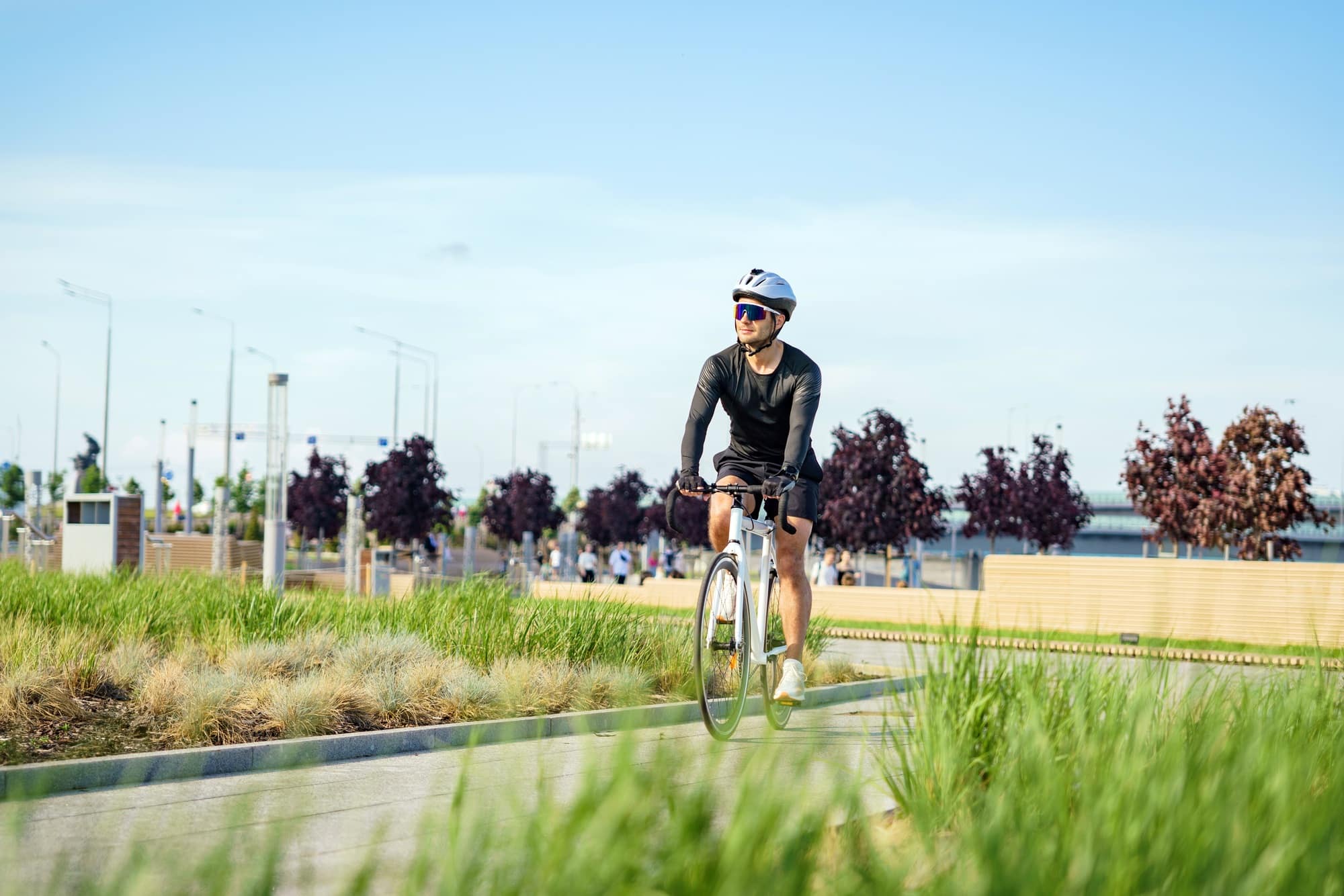 A cyclist in a black outfit rides through a green park on a sunny day, surrounded by nature and city