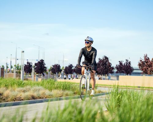 A cyclist in a black outfit rides through a green park on a sunny day, surrounded by nature and city